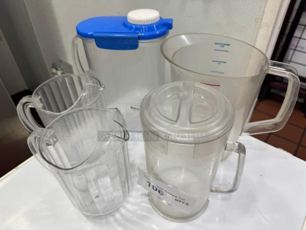 Set of 5 pitchers, various styles, clear plastic, one with blue lid, measuring marks visible, auction tag labeled "106 QTY 5."
