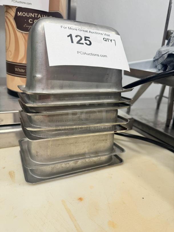 Stainless steel food pan BIDX7, set of seven, used condition, stacked with auction tag "125 QTY 7" visible, showing minor wear.