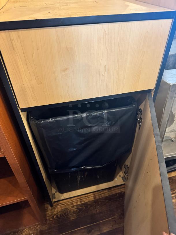 Wood finish gallon waste receptacle with hinged door, black trim, used condition. Inner black bag visible. No brand markings.