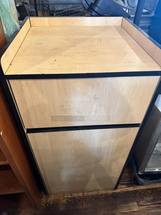 Wooden gallon waste receptacle with black trim, natural finish, and visible wear. Rectangular shape, suitable for indoor use.