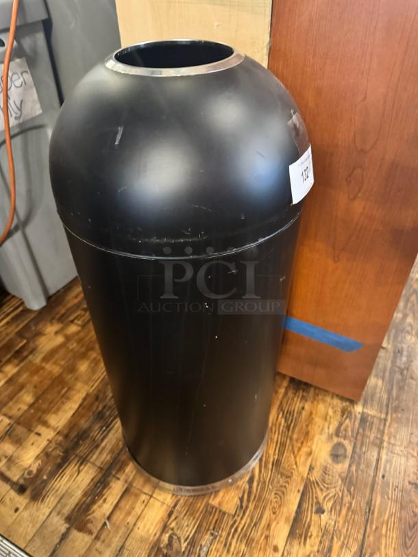 Black open top trash can with cylindrical shape, minor wear, placed on wooden floor. Label marked "132".