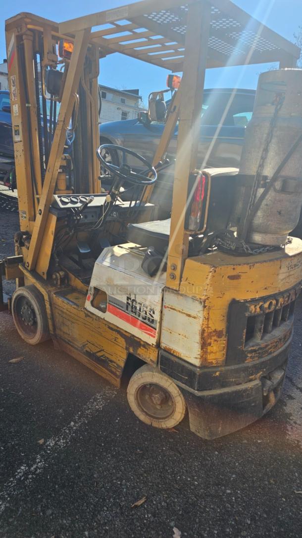 KOMATSU FG 15ST-2 forklift, yellow, good condition, visible wear. Label indicates model. Includes safety features, gas tank.