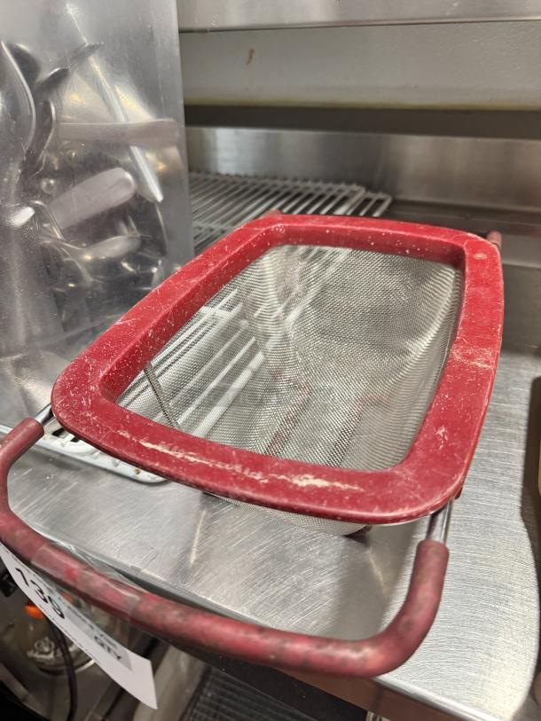 Red-handled colander/strainer with metal mesh, visible wear, suitable for kitchen use.