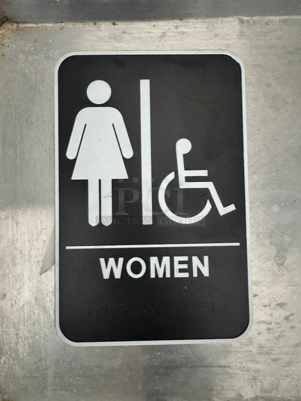 Women's restroom sign with handicap symbol, black background, white icons and text, mounted on a metallic surface.
