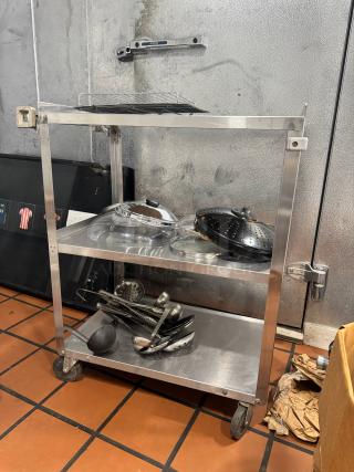 Stainless steel utility cart with three shelves, holding kitchen utensils. In good condition with visible caster wheels for mobility.