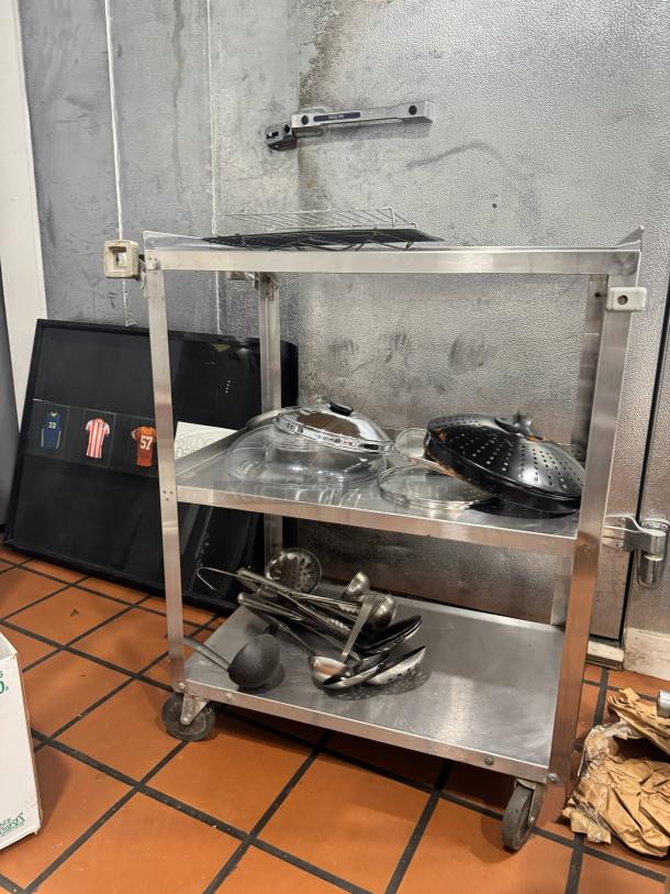 Stainless steel utility cart with two shelves, loaded with kitchen utensils, on wheels for easy mobility. Good condition.