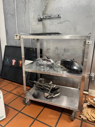 Stainless steel utility cart with two shelves, filled with kitchen utensils and lids. Features wheels for mobility. Good condition.