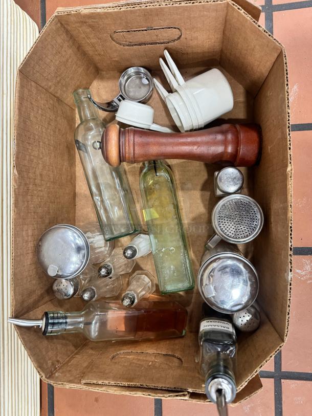 Box of miscellaneous kitchenware including glass bottles, wooden pepper mill, measuring cups, and stainless steel shakers.