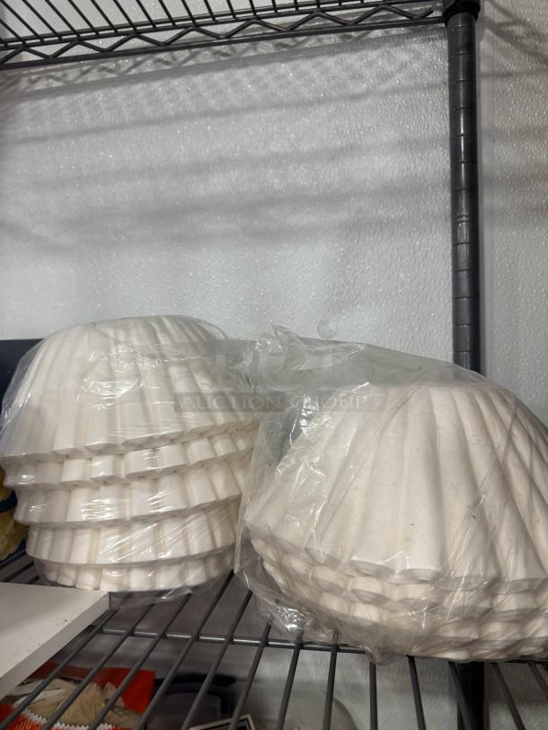 Stacks of white coffee filters on a metal shelf, some in plastic wrap, indicating unused condition.