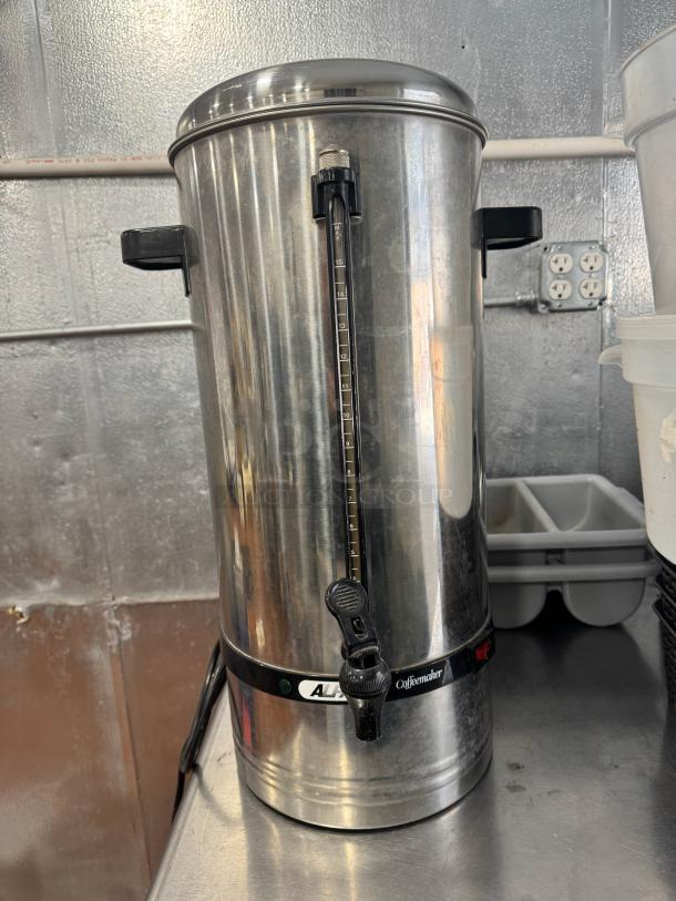 Stainless steel coffee urn/percolator with a black spout and handles, built-in level gauge, and "All American Coffeemaker" label.