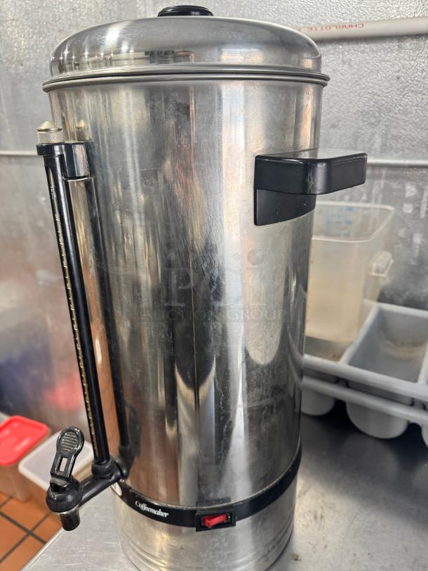 Stainless steel coffee urn/percolator with black handles, spigot, and indicator light. Visible "Coffeemaker" label.