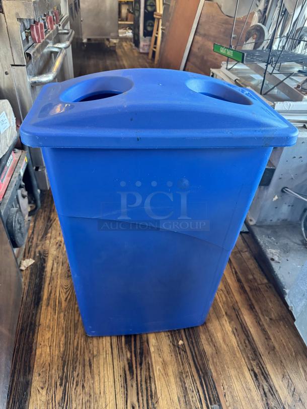23-gallon blue slim recycling bin, dual circular openings on lid, in used condition, shown on wooden floor.