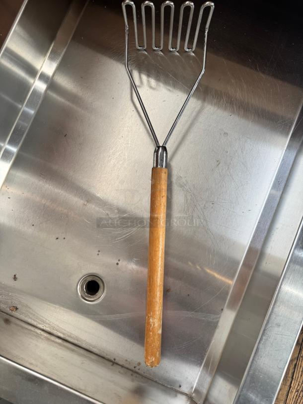 24" potato masher with wooden handle, metal mashing surface, used condition, shown resting in a metal sink.