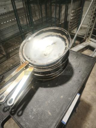 Stack of 12" stainless clad frying pans with heat-worn surfaces and metal handles, indicating heavy use.