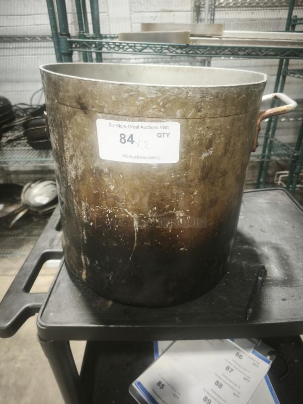 Extra large aluminum stock pot with a handle, visible wear, auction label noting lot 84, ideal for high-volume cooking.