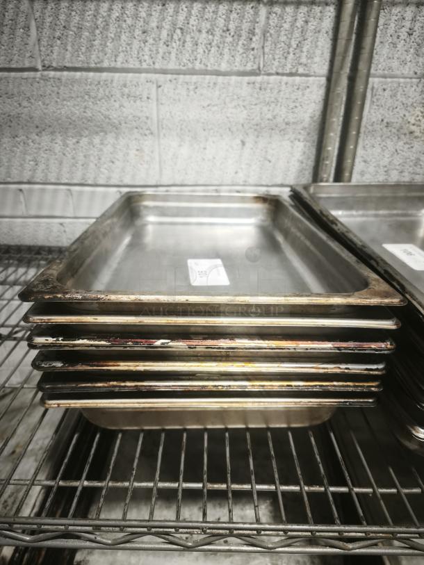 Full-size rectangular food pans stacked on a metal shelf, showing wear and light scratches, visible auction tag.
