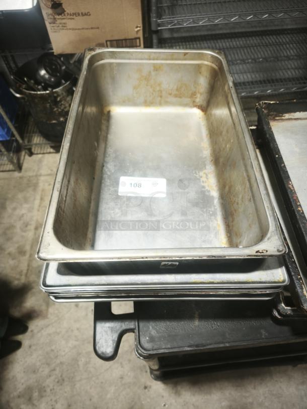 Stainless steel steam table pans, full size, 4" deep, showing signs of use with discoloration, stacked in a storage area.