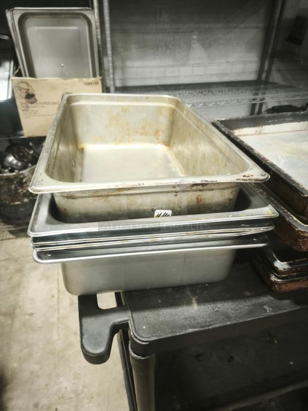 Stainless steel full-size steam table pans, 4" deep, showing signs of use, stacked on a cart in a storage area.