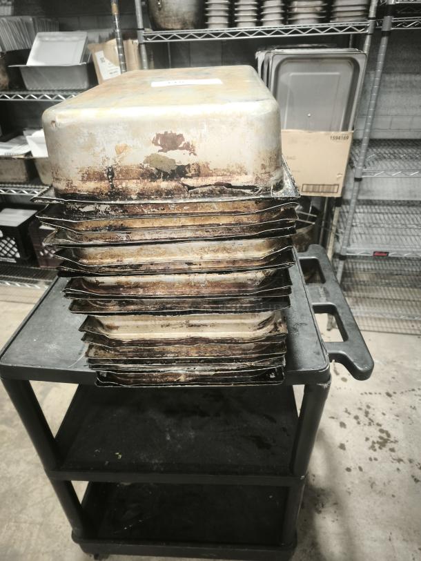 Stack of stainless steel steam table pans, full size, 4" deep, with visible wear and discoloration, on a cart.