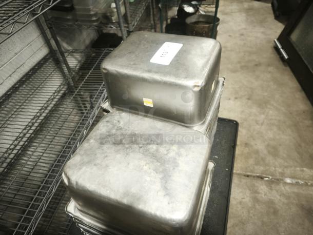 Stainless steel steam table pans, half full size, 6" deep, used condition with visible wear. Stacked with labels.