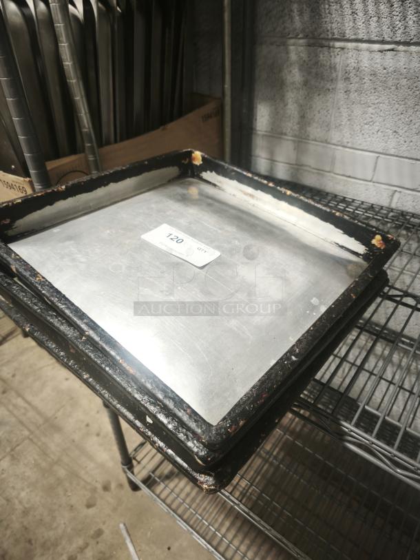 Stack of rectangular stainless steel trays with worn edges on a metal shelf. Label shows "120".