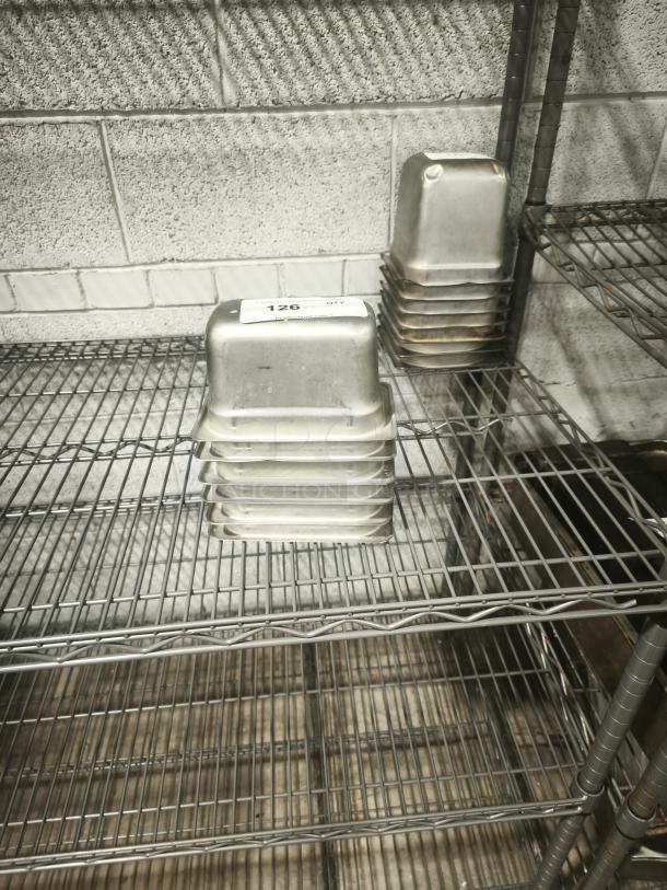 Stack of 1/9 size 4" deep stainless steel food pans on metal shelving, showing light wear. Label with "126 QTY" visible.
