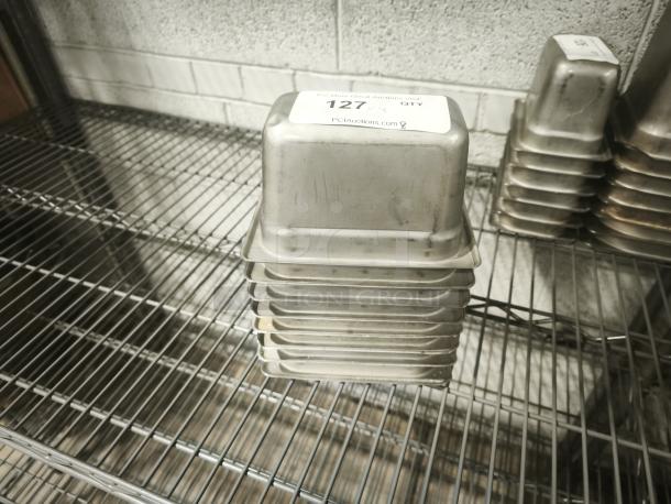 Stack of stainless steel 1/9 size, 4" deep food pans on metal shelving, tag displaying auction information on top pan.