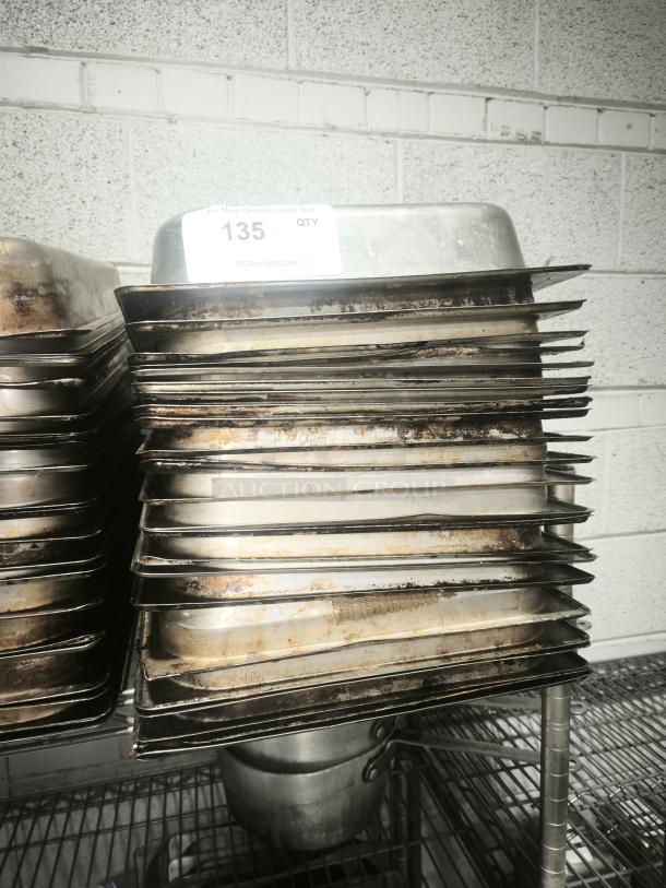 Stack of full-size 4" deep food pans on metal shelf, showing signs of wear. Auction tag reads "135 QTY" from PCI Auctions.