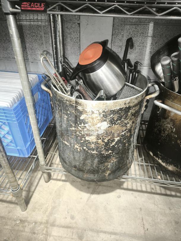 Large, heavily worn metal pan with cooking utensils inside, including a jug with an orange lid. Stored on a metal shelf.