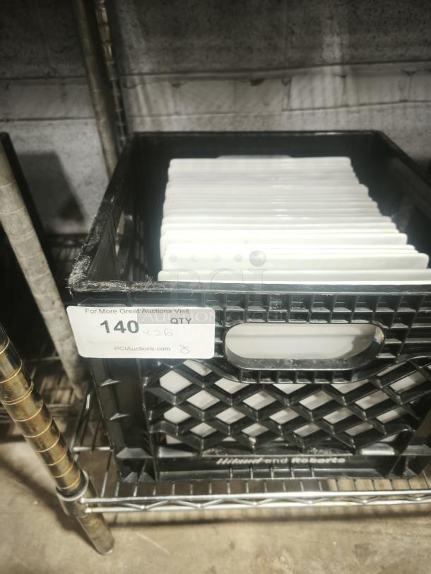 9" bright white square porcelain plates stacked in a crate, quantity 26 label visible, stored on a metal shelf.