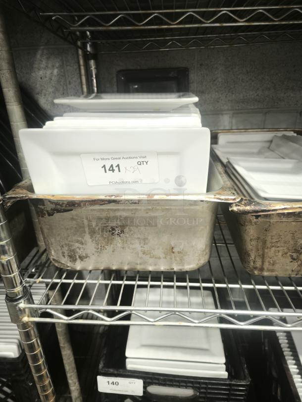 Stacked 9" bright white square porcelain plates on a metal shelf, numbered tag indicating quantity for auction.