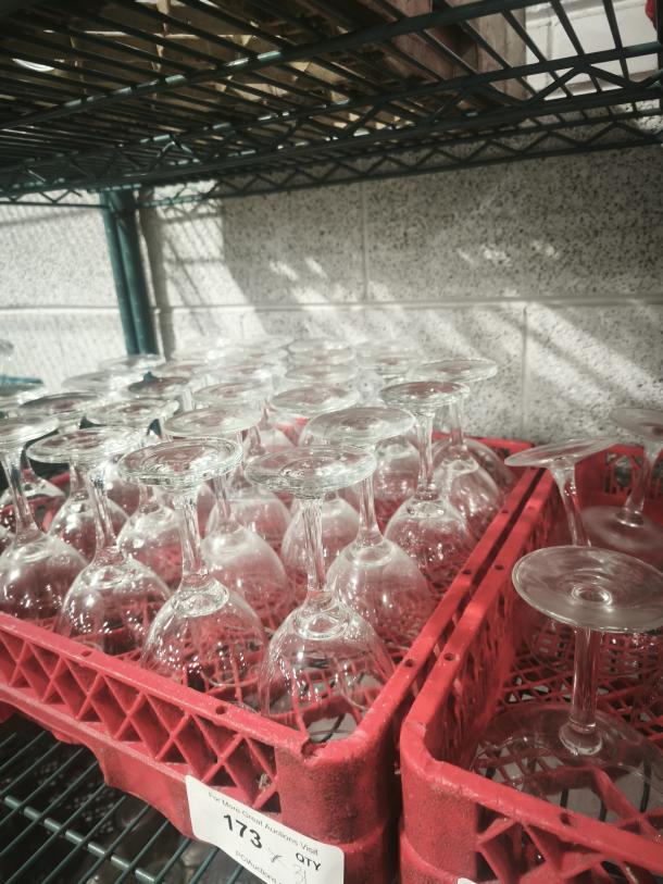 Clear wine glasses stored upside down in red racks on a wire shelf. Racks labeled with auction details. Glasses appear unused.