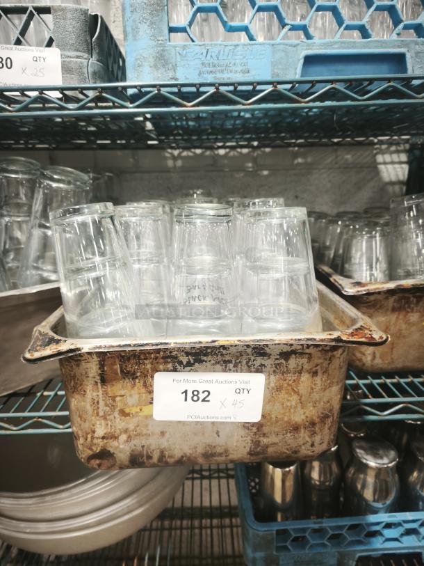 Vintage restaurant glass tumblers in used condition, stacked in a rusty metal container, labeled "182 QTY 45".