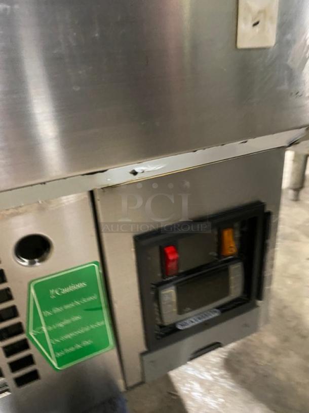 Commercial stainless steel refrigerated bakery display case with straight glass, multi-tier shelving, and rear access sliding doors. Visible wear and control panel.