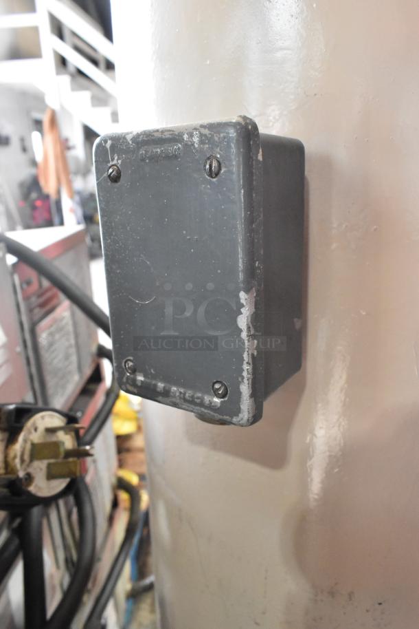 A grey metal electrical junction box attached to a painted pole, showing signs of wear and slight chipping.