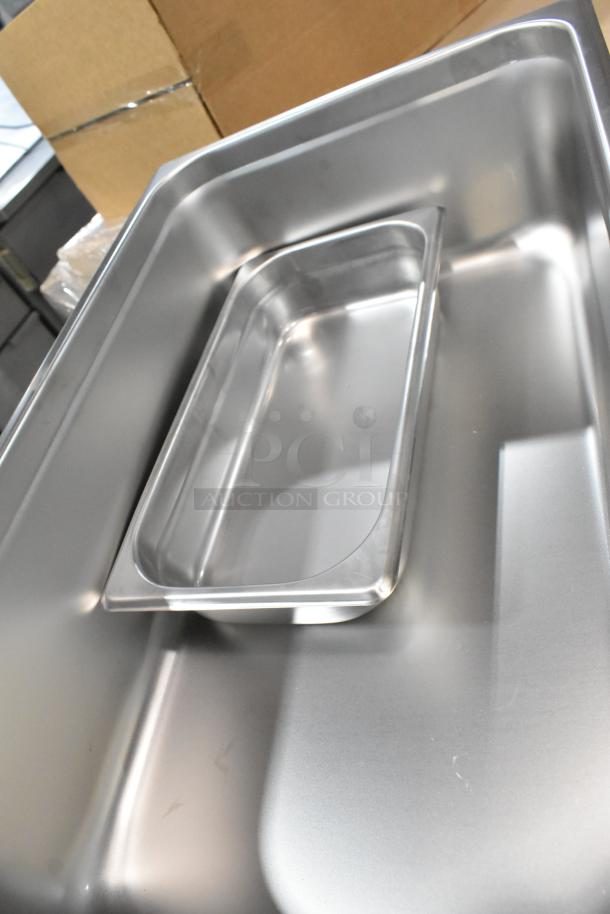 Brand new stainless steel pans with lids featuring a smooth finish, part of a 42-piece set for auction.