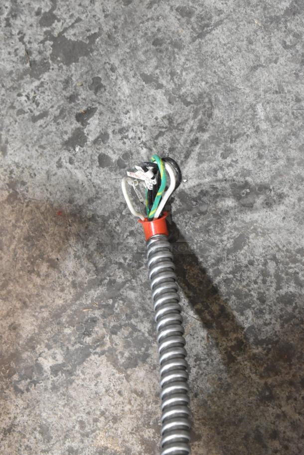Coiled metal conduit on gray concrete floor, with exposed wiring at the end, labeled for identification.