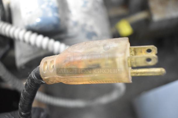 Close-up of a worn electric plug with visible prongs and frayed wiring on a blurred industrial background.