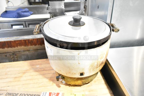 Metal countertop rice cooker with stainless steel lid, showing signs of wear and rust. Features side handles and steam vent.