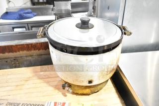 Metal countertop rice cooker with stainless steel lid, showing signs of wear and rust. Features side handles and steam vent.