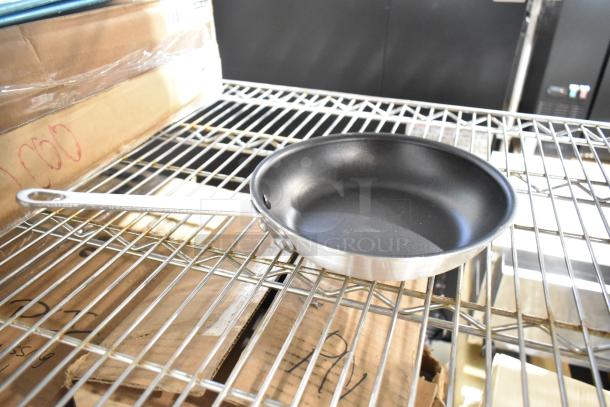 Brand new stainless steel frying pan with a black non-stick interior, resting on a metal rack among boxed items.