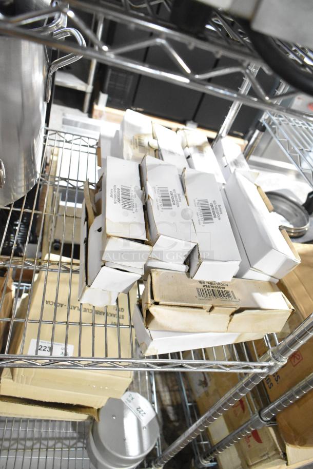 Boxes of 24 new scratch and dent Choice Bethany 8 3/8" 18/0 stainless steel tablespoons on metal shelves.