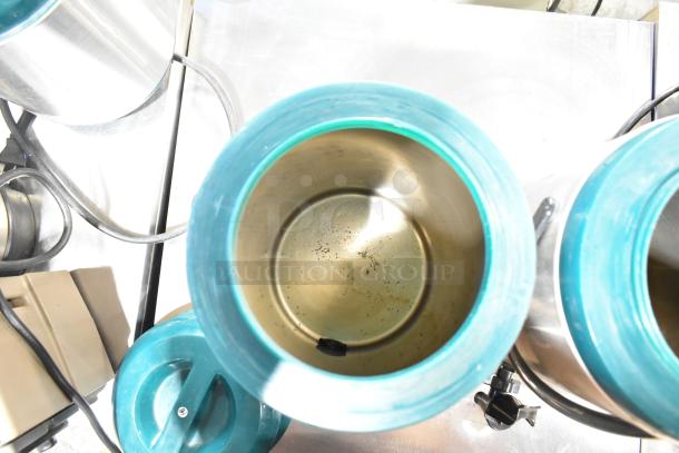 Two stainless steel beverage dispensers with teal lids, featuring spouts and power cords, shown from above on a metal surface.