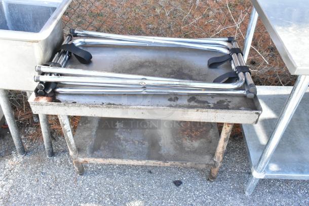 Metal table with under shelf, weathered condition, visible rust and discoloration, located outdoors with nearby metal items.