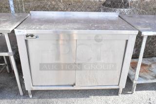 Stainless steel table with metal under shelves and sliding doors, some surface wear visible, industrial design.