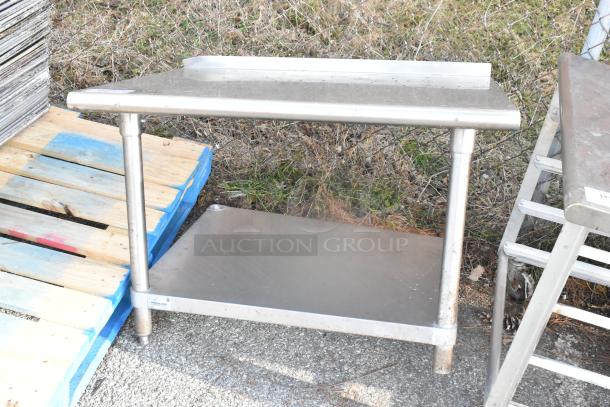 Stainless steel table with metal under shelf, outdoor setting, minor wear, sturdy construction, ideal for commercial use.