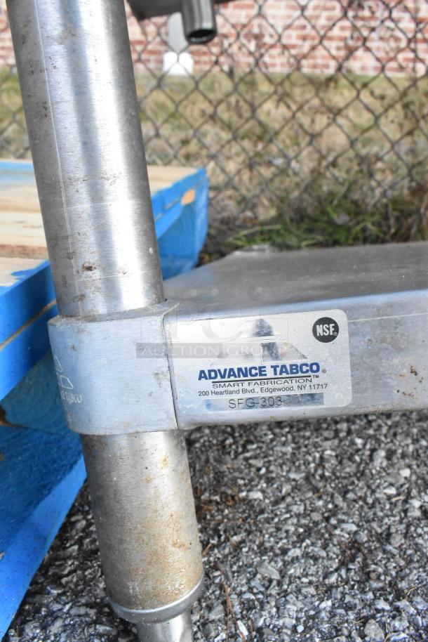 Stainless steel table with metal under shelf, labeled "Advance Tabco SFG-303" with NSF certification, featuring sturdy construction.