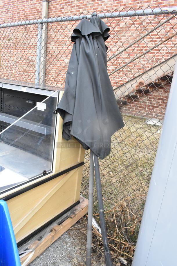 Two patio umbrellas, black fabric, steel poles, leaning against a chain-link fence. Gently used, with minor wear.