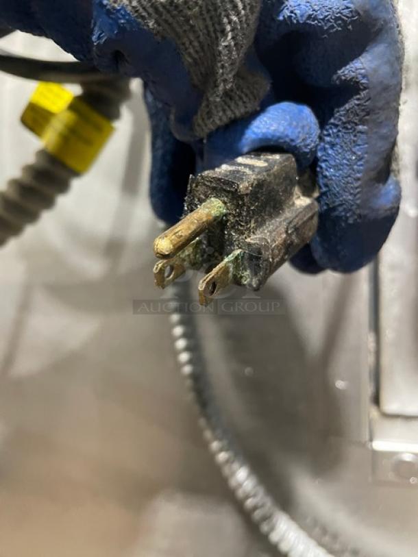 A close-up of a worn electrical plug with corrosion, held by a gloved hand. Background shows part of a cable.