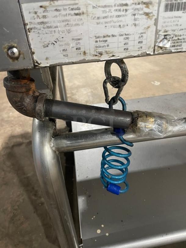 WoodStone griddle connection detail, showing gas line, coiled safety cable, and partially visible label on a stainless steel stand.